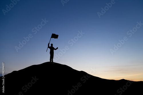 Silhouette of businessman holding a flag on top mountain, sky and sun light background. Business success and goal concept. © cofficevit