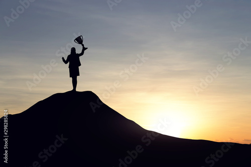 Silhouette of businesswoman with a championship cup celebrate success on top mountain, sky and sun light background.