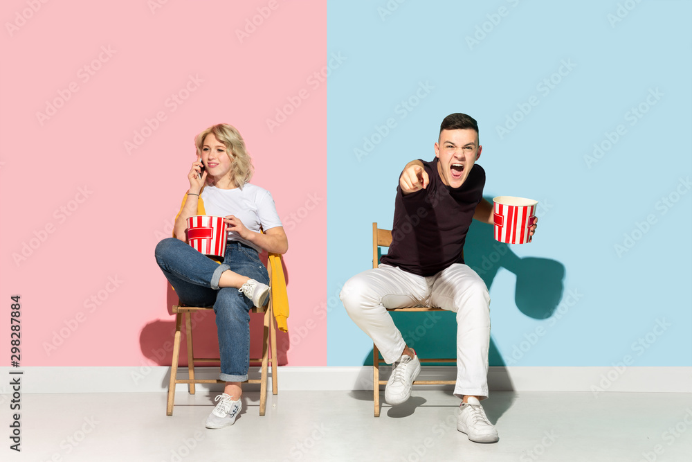 Obraz premium Young emotional man and woman in bright casual clothes posing on pink and blue background. Concept of human emotions, facial expession, relations, ad. Beautiful couple watching cinema with popcorn.