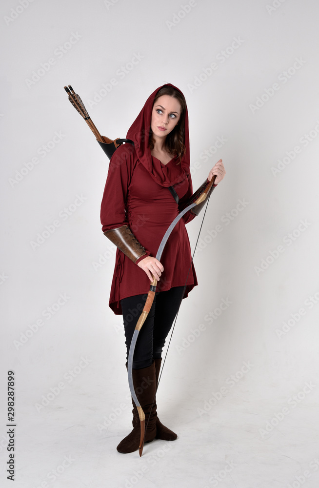 full length portrait of a brunette girl wearing a red fantasy tunic with hood, holding a bow and arrow. Standing pose on a white studio background. full length portrait of a brunette girl wearing a red fantasy tunic with hood, holding a bow and arrow. Standing pose on a white studio background.