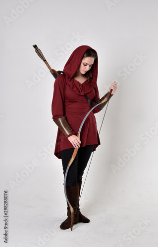 фотографія full length portrait of a brunette girl wearing a red fantasy tunic with hood, holding a bow and arrow
