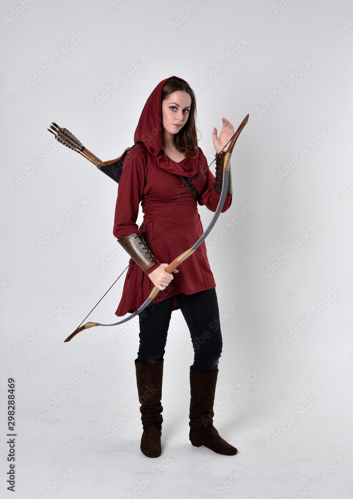 full length portrait of a brunette girl wearing a red fantasy tunic with hood, holding a bow and arrow. Standing pose on a white studio background. full length portrait of a brunette girl wearing a red fantasy tunic with hood, holding a bow and arrow. Standing pose on a white studio background.