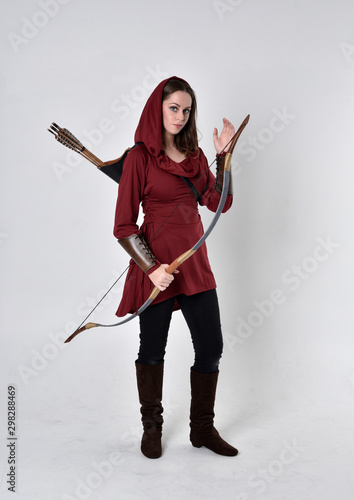 full length portrait of a brunette girl wearing a red fantasy tunic with hood, holding a bow and arrow. Standing pose on a white studio background.