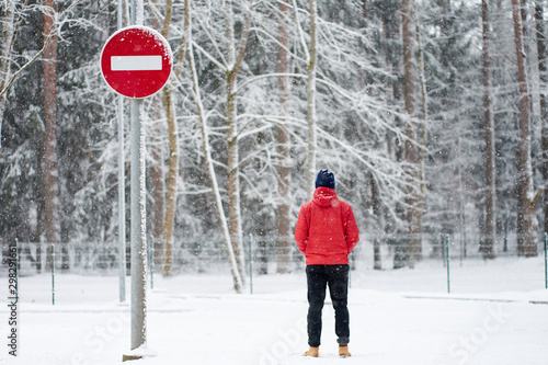 Man wearing red winter jacket standing near no entrans road sign during snowfall