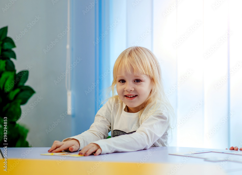 Little girl in white clothes looking in the camera and smiling. Adorable blonde kid is Sitting at the table. Closeup. Portrait Little girl in white clothes looking in the camera and smiling. Adorable blonde kid is Sitting at the table. Closeup. Portrait