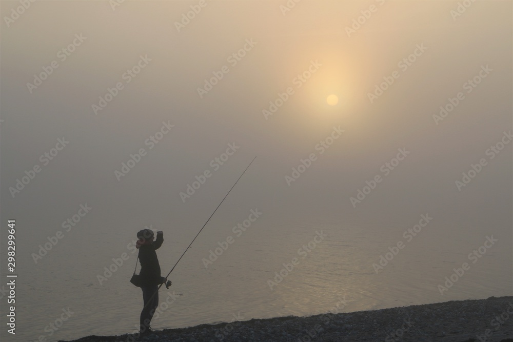 silhouette of a female angler with spinning rod in the fog by the sea