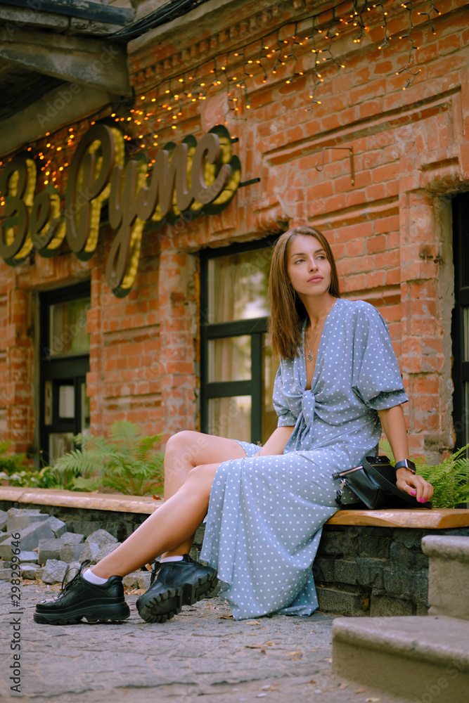 Blonde girl in long blue dress and small black handbag on her shoulder is posing sitting sideways on a parapet. Fashion and style. Full length shot. Blonde girl in long blue dress and small black handbag on her shoulder is posing sitting sideways on a parapet. Fashion and style. Full length shot.