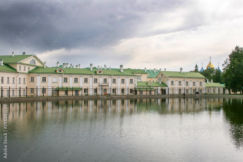 A long two-story house with a green roof, located behind a fence on the pond on an overcast day. In the distance are the domes of the Holy Trinity St. Sergius Lavra, the city of Sergiyev Posad