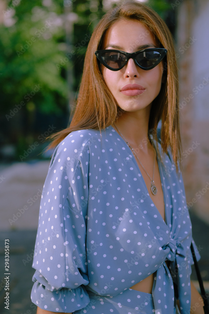 Blonde girl in long blue dress, sunglasses and a small black handbag on her shoulder is walking alone in the city. Fashion and style. Close-up shot. Blonde girl in long blue dress, sunglasses and a small black handbag on her shoulder is walking alone in the city. Fashion and style. Close-up shot.