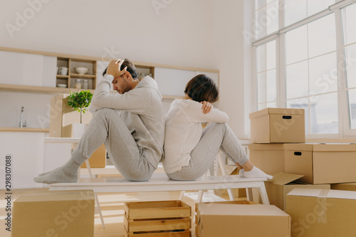 Photo of stressful couple get divorse, leave house, start living seperately, sit backs to each other, pose in kitchen, cardboard boxes with belongings around, focused down with sad expressions photo