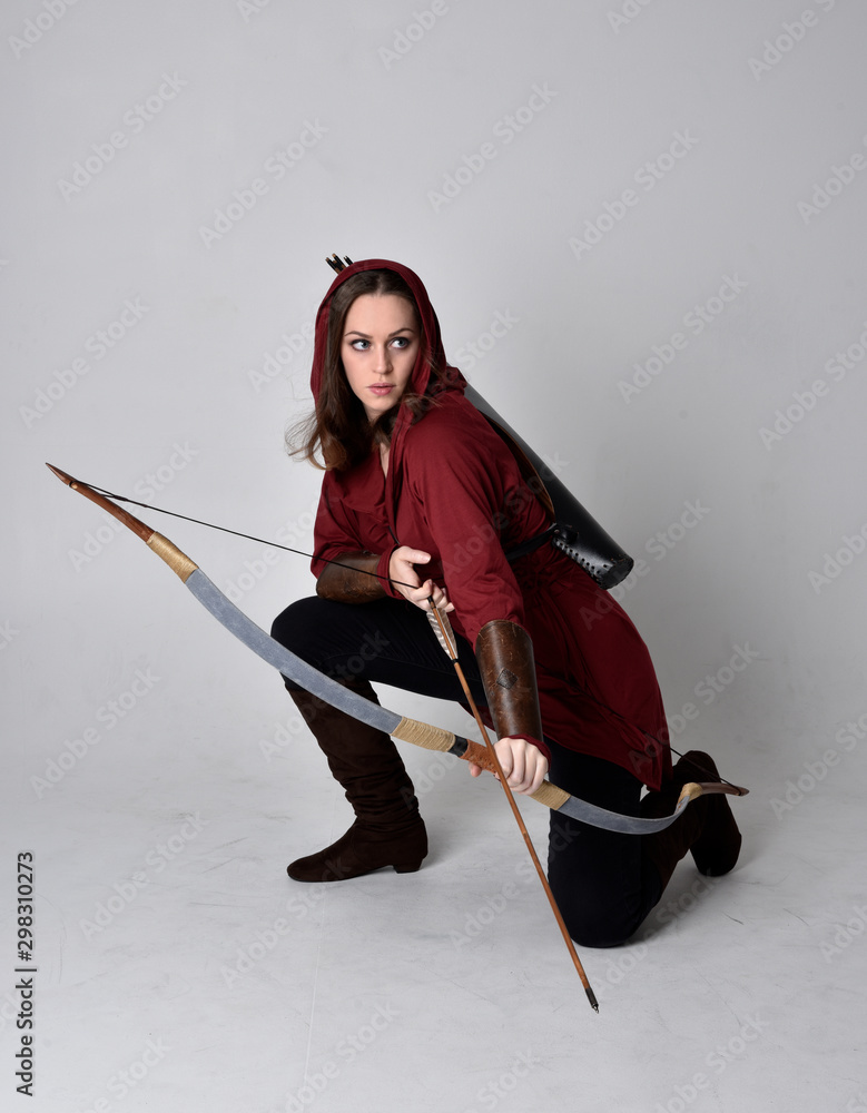 Φωτογραφία full length portrait of a brunette girl wearing a red fantasy tunic with hood, holding a bow and arrow