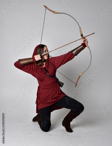 Fototapet full length portrait of a brunette girl wearing a red fantasy tunic with hood, holding a bow and arrow