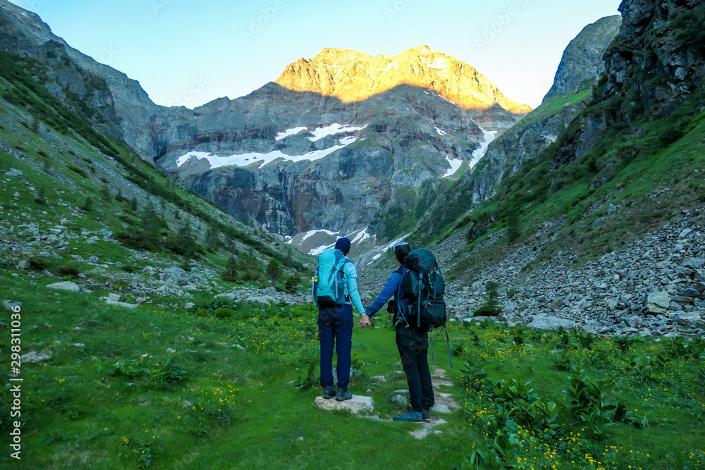A couple standing on a meadow with the view on Schladming Alps, partially still covered with snow. Spring slowly reaching the tallest parts of the mountains. Soft sunrise on the mountain peaks.