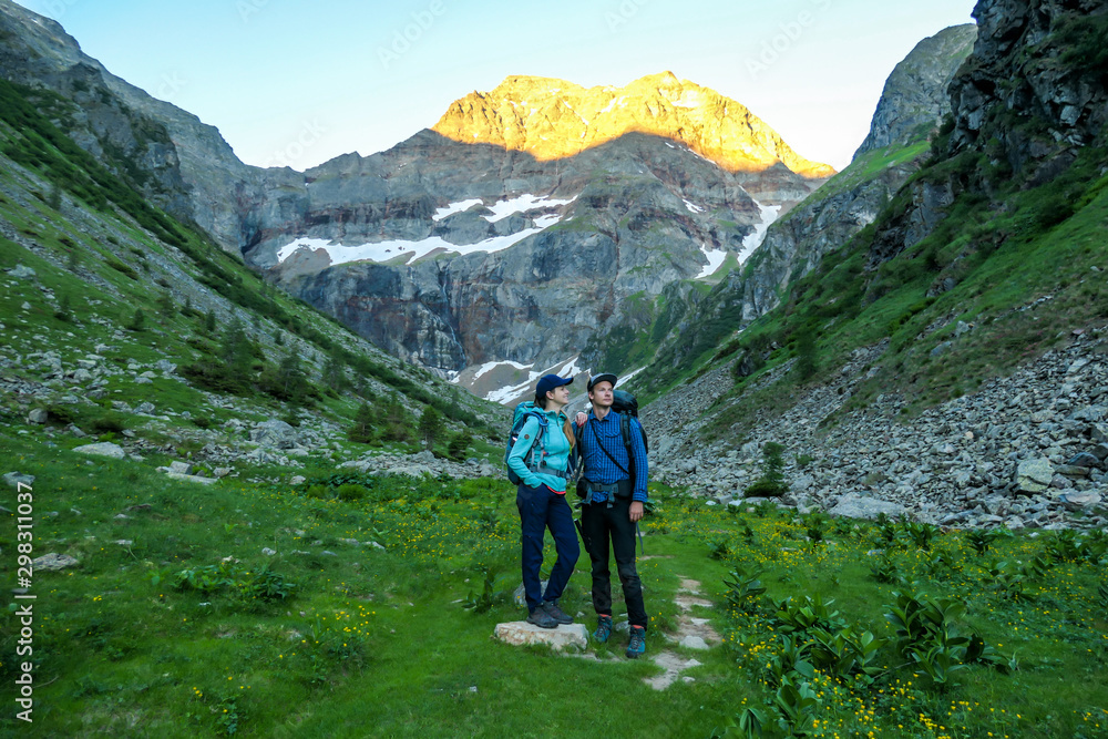 A couple standing on a meadow with the view on Schladming Alps, partially still covered with snow. Spring slowly reaching the tallest parts of the mountains. Soft sunrise on the mountain peaks.
