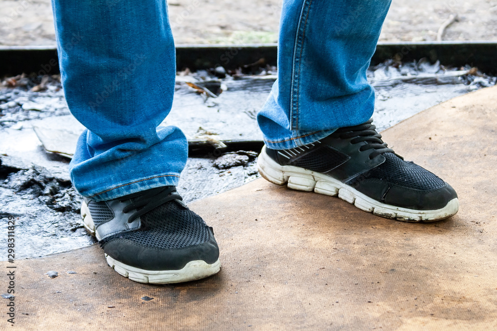 Men's legs in blue jeans and black sneakers with white soles stand on a dirty oiled plywood board. Dirty car master workplace Men's legs in blue jeans and black sneakers with white soles stand on a dirty oiled plywood board. Dirty car master workplace