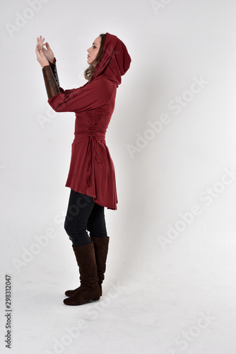 full length portrait of a brunette girl wearing a red fantasy tunic with hood. Standing pose in side profile on a white studio background. photo