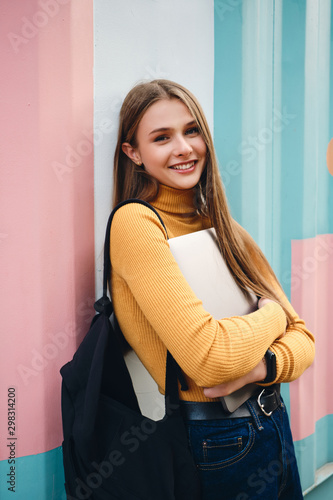 Pretty joyful casual student girl with laptop happily looking in camera over colorful background outdoor