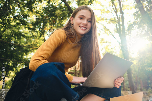 Beautiful smiling student girl joyfully looking in camera studying on laptop in park