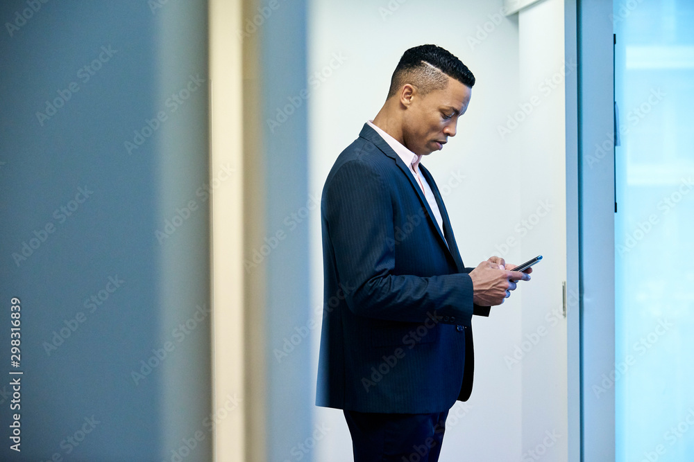 Mid adult businessman text messaging on smart phone while standing in hotel lobby Mid adult businessman text messaging on smart phone while standing in hotel lobby