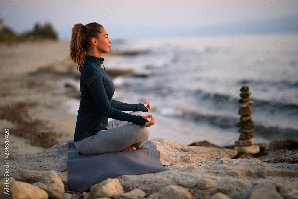 Young woman meditating in lotus position on a rock at the beach.