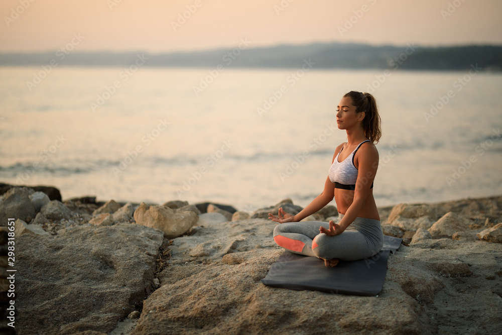 Athletic woman meditating with eyes closed on a rock by the sea.