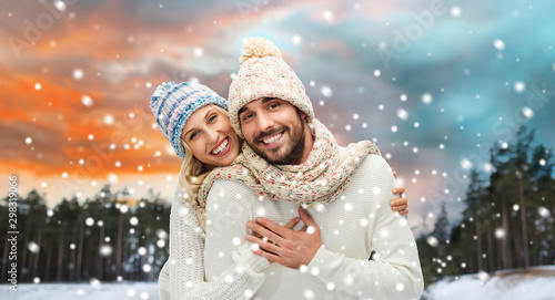 couple, christmas and holidays concept - smiling man and woman in hats and scarf hugging over winter forest background