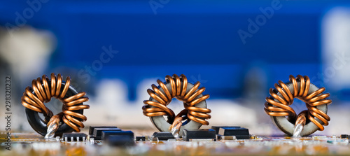 Panoramic electrotechnical background. Toroidal inductors with beautiful copper wire winding. Electronic components as coils or transistors on circuit board detail. Copy space on a blue blurry device.