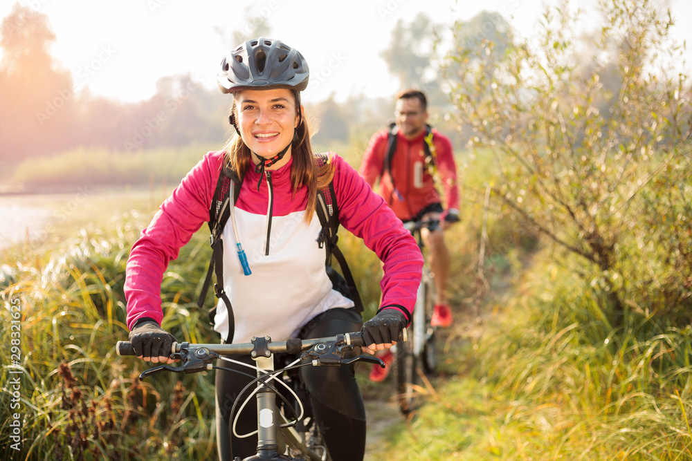 Beautiful happy young woman riding mountain bike over a meadow by the lake or river in the early morning. Young man blurred in the background. Healthy and active sporty lifestyle