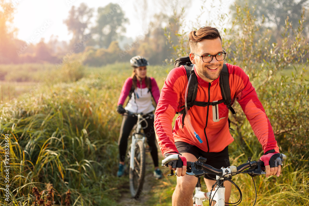 Obraz premium Beautiful happy young man riding mountain bike over a meadow by the lake or river in the early morning. Young woman blurred in the background. Healthy and active sporty lifestyle
