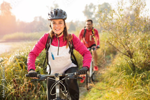 Obraz na płótnie Beautiful happy young woman riding mountain bike over a meadow by the lake or river in the early morning. Young man blurred in the background. Healthy and active sporty lifestyle w salonie