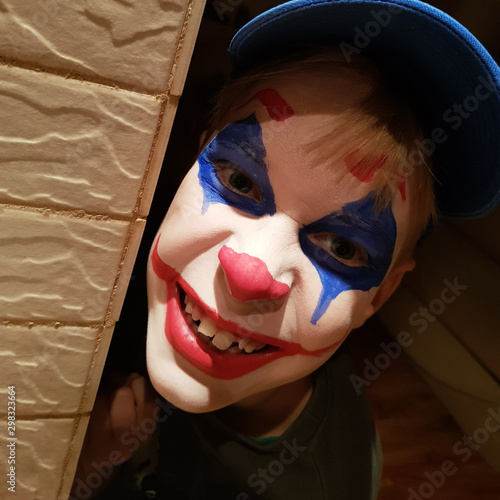 A cheerful boy with a clown mask painted on his face peeks around the corner. Body painting and makeup for a school carnival or halloween celebration.