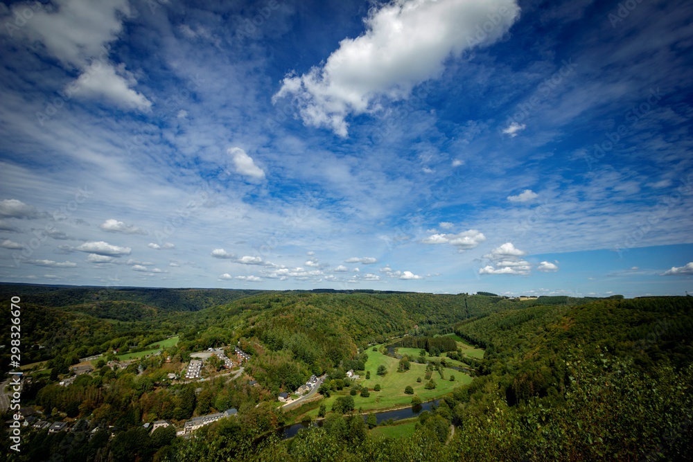 A landscape photograph of the area of Laforêt in the ardennes in Belgium with a blue sky and lots of forest. There is a village and the Semois river flowing through the woods.