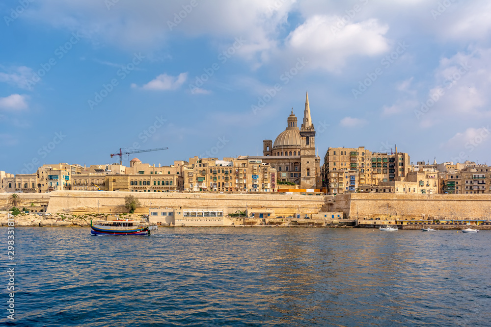 Coastal landscape of Valletta at sunny summer day, with boat drifting in the foreground. Maltese Valletta skyline with church of Our Lady of Mount Carmel and St Paul's Pro-Cathedral.