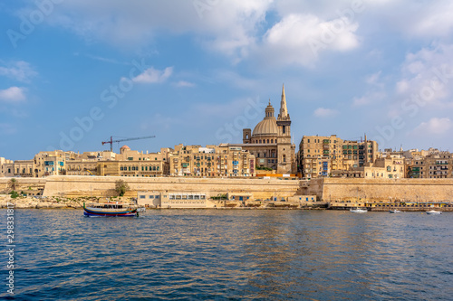 Coastal landscape of Valletta at sunny summer day, with boat drifting in the foreground. Maltese Valletta skyline with church of Our Lady of Mount Carmel and St Paul's Pro-Cathedral.
