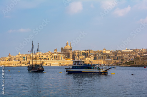 Coastal landscape of Valletta at sunny summer day, with modern boat and medieval ship in the foreground. Maltese Valletta skyline with church of Our Lady of Mount Carmel and St Paul's Pro-Cathedral.