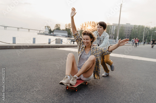 Fototapeta Naklejka Na Ścianę i Meble -  Cheerful son riding his mother on a skateboard and laughing. Single mother spending time with her son outdoors