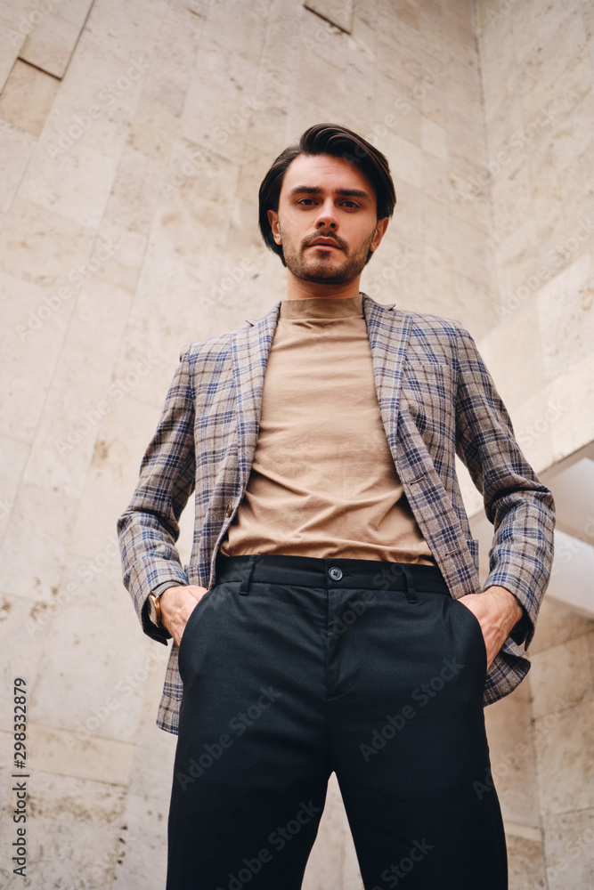 Obraz premium Young handsome man in checkered jacket holding hands in pockets confidently looking in camera outdoor