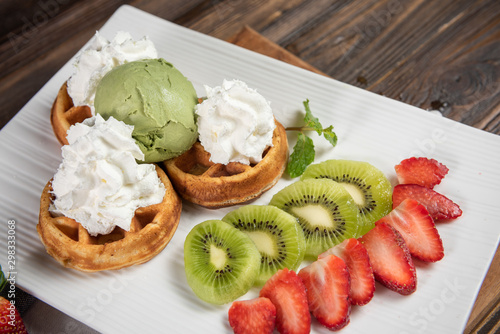 The green tea waffle, topping with whipped cream and green tea ice cream on a white plate. Decorated with fresh fruit as strawberry, kiwi and peppermint. Dark brown real wood chopping and background.