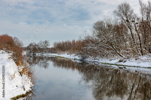 Small river in winter at sunny day against cloudy sky. Country winter landscape