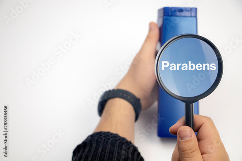 Chemical components on the shampoo label: parabens . A hand holds a blue jar and a magnifier, where the harmful ingredients of a detergent are written in close up. photo