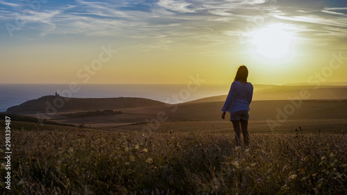 Lone silhouette female stands in a wide open landscape facing the sea as the sun shines in the distance