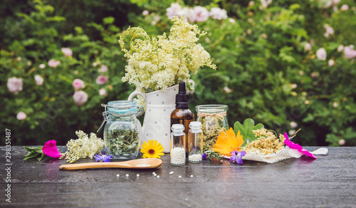 Foto Homeopathic medicine pills on wood table, globules in jars and on wood spoon, decorated with fresh and dry various herbal plants outdoors in summer, greenery background