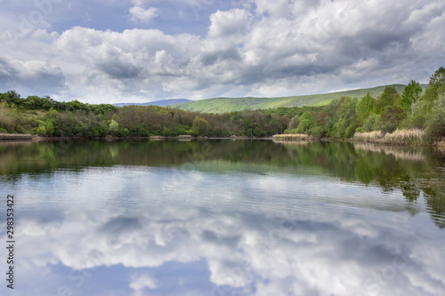Fototapeta Naklejka Na Ścianę i Meble -  Beautiful, sunlit, vivid green trees in a forest during early spring around Sukovo lake reflecting the trees and cloudy sky in a calm water