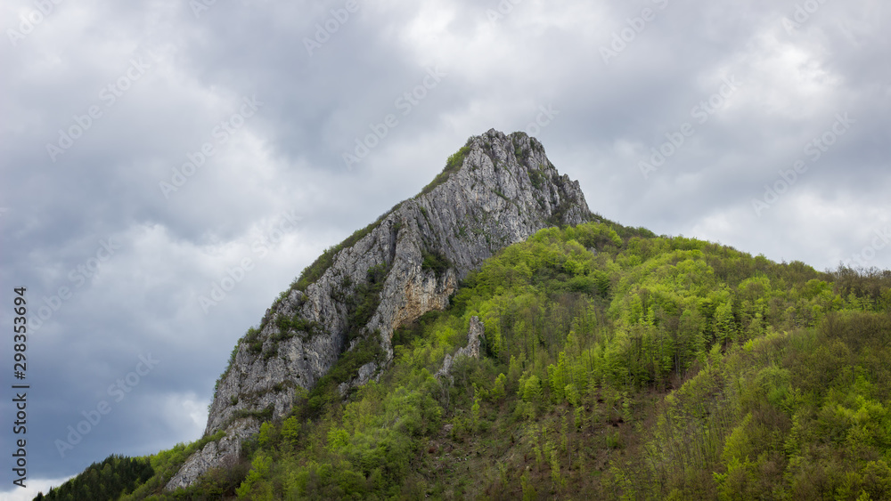 Impressive, rocky, pointy summit Asenovo kale, dramatic cloudy sky and beautiful sunlit, vivid green forest during early spring