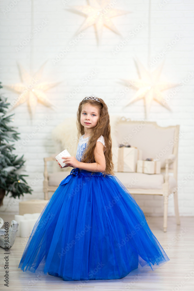 Cute little girl in a magnificent blue dress holding an artificial candle in her hands for home decoration on Christmas holidays. Child-friendly scenery