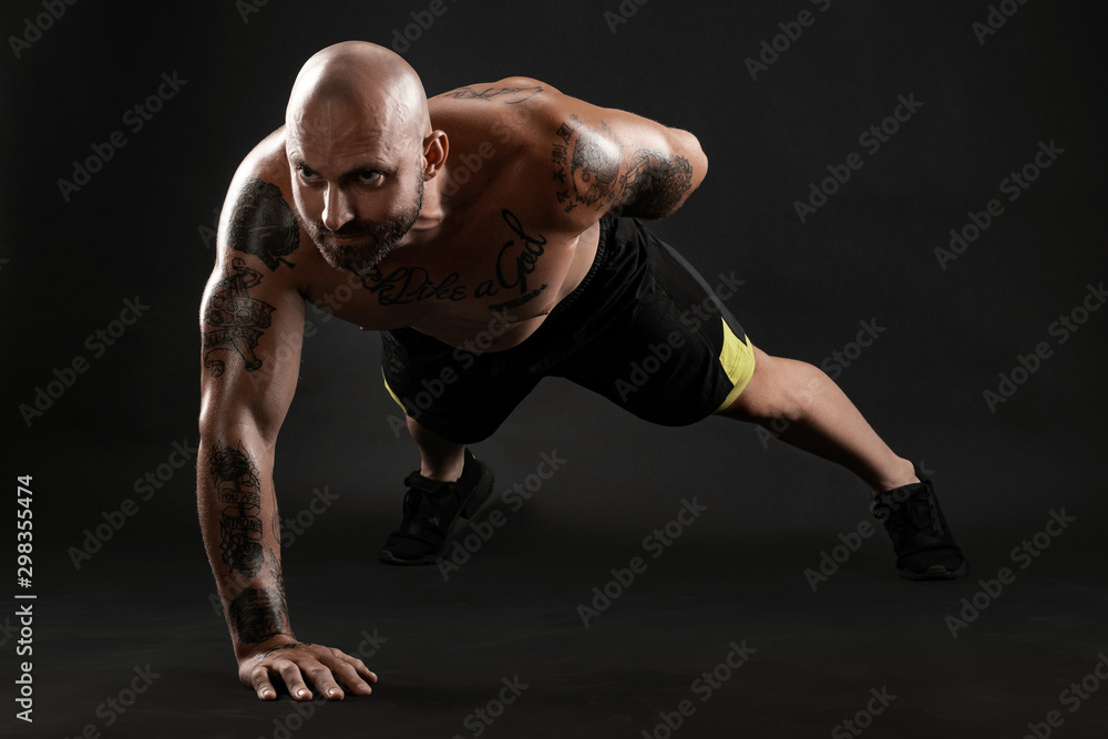 Athletic bald, tattooed man in black shorts and sneakers is posing against a black background. Close-up portrait. Athletic bald, tattooed man in black shorts and sneakers is posing against a black background. Close-up portrait.