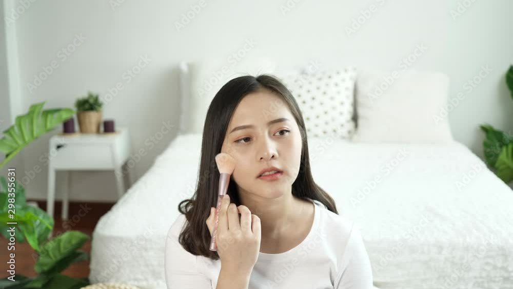 Asian woman makes makeup oneself. Young woman makes blush on the face using makeup brush sitting in the bedroom in the morning there was sunshine from the window. Beauty makeup concept. Slow motion Asian woman makes makeup oneself. Young woman makes blush on the face using makeup brush sitting in the bedroom in the morning there was sunshine from the window. Beauty makeup concept. Slow motion