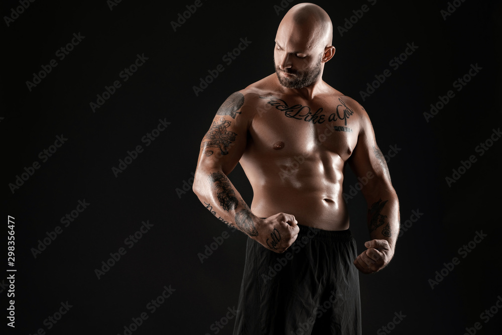 Athletic bald, bearded, tattooed man in black shorts is posing against a black background. Close-up portrait.