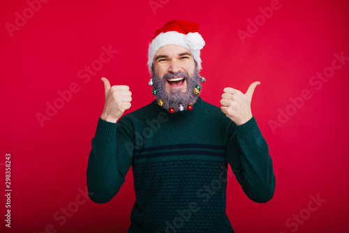 Happy man with white decorated beard wearing santa claus hat and showing thumbs up photo