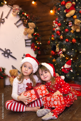 Little sisters girls in red pajamas decorate Christmas tree and open gifts in living room. Family with kids celebrating christmas at home. Holiday gifts for baby. Merry Christmas and happy holidays. 